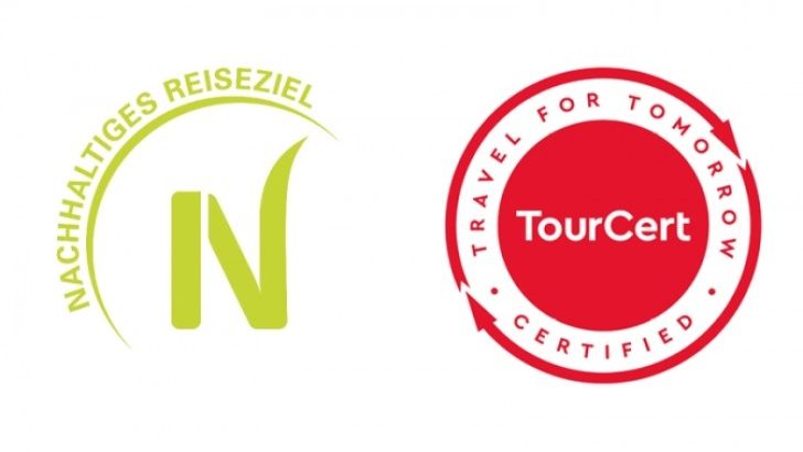 Das Bild zeigt zwei nebeneinander platzierte Zertifizierungslogos. Links befindet sich ein grün gehaltenes „N“-Logo mit dem halbkreisförmigen Schriftzug „NACHHALTIGES REISEZIEL“. Rechts daneben steht ein rundes rotes Siegel mit dem Schriftzug „TourCert“ in der Mitte und dem umlaufenden Text „TRAVEL FOR TOMORROW – CERTIFIED“, was auf eine Nachhaltigkeitszertifizierung im Tourismussektor hinweist.