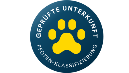 Das Bild zeigt ein rundes blaues Siegel mit einem großen gelben Pfotenabdruck in der Mitte. Am oberen Rand steht in weißen Großbuchstaben der Text „GEPRÜFTE UNTERKUNFT“. Am unteren Rand befindet sich der Schriftzug „TIERFREUNDLICHKEIT GEPRÜFT“, was darauf hinweist, dass die Unterkunft offiziell als tierfreundlich zertifiziert wurde.
