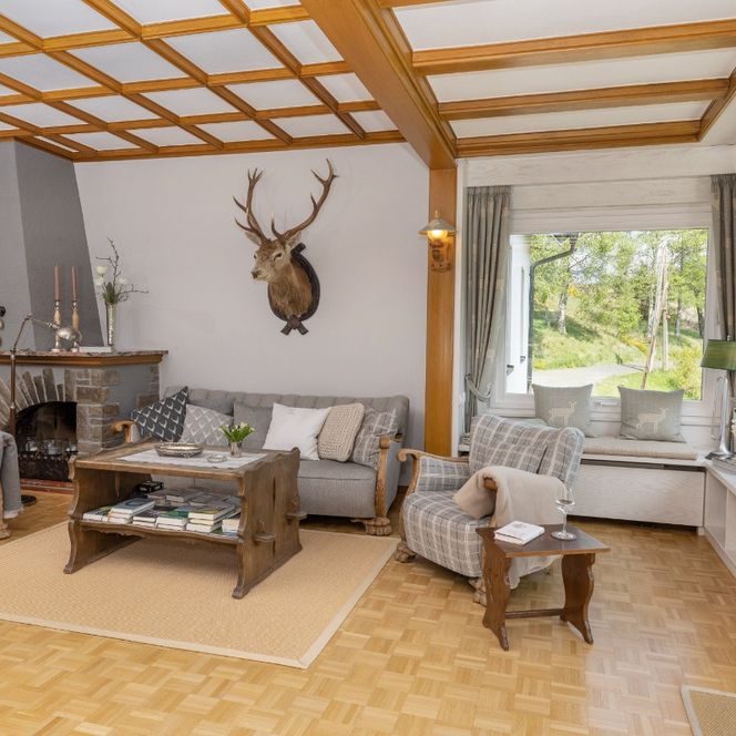 Das Bild zeigt ein großzügiges, helles Wohnzimmer mit mehreren bequemen Sesseln und Sofas, die um einen niedrigen Holztisch gruppiert sind. An der Wand hängen ein Hirschgeweih und stilvolle Dekorationen, während große Fenster viel Tageslicht hereinlassen und den Blick ins Grüne freigeben. Der Raum wirkt einladend, elegant und hat eine warme, ländliche Atmosphäre.