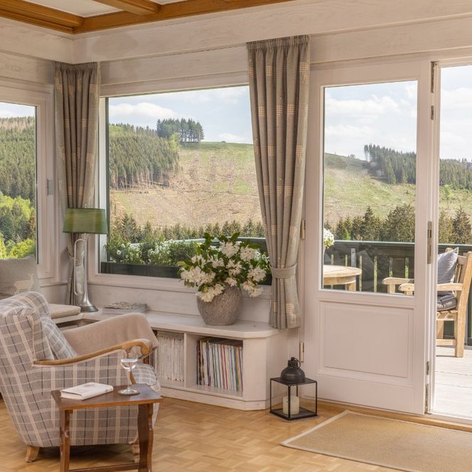 Das Bild zeigt einen hellen Wohnbereich mit großen Fenstern, durch die viel Tageslicht hereinfällt und einen weiten Blick auf eine grüne Hügellandschaft freigibt. Vor den Fenstern stehen gemütliche Sessel, Dekorationen und ein kleiner Tisch, die eine einladende, wohnliche Atmosphäre schaffen. Rechts führt eine geöffnete Terrassentür auf einen Balkon, der den Raum optisch mit der Natur verbindet.