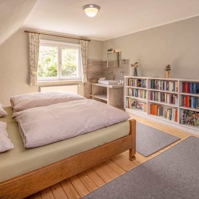 Das Bild zeigt ein gemütlich eingerichtetes Schlafzimmer mit einem großen Holzbett, das mit hellen Bettdecken bezogen ist. An der rechten Wand steht ein farbenfrohes Regal voller Bücher, daneben ein kleiner Schreibtisch unter einem Fenster, durch das warmes Tageslicht hereinfällt. Der Raum wirkt hell, einladend und bietet eine angenehme Mischung aus Schlaf- und Arbeitsbereich.