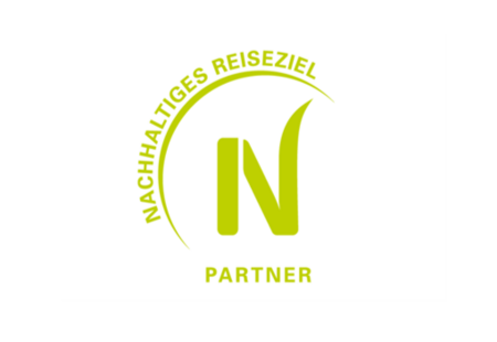 Das Bild zeigt ein Logo mit einem großen grünen Buchstaben „N“ in der Mitte. Darüber verläuft ein halbkreisförmiger Schriftzug „NACHHALTIGES REISEZIEL“, ebenfalls in Grün. Unter dem Buchstaben steht das Wort „PARTNER“, wodurch das Logo wie ein Siegel für nachhaltigen Tourismus wirkt.