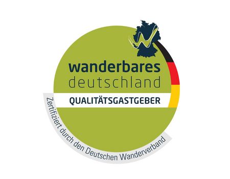 Das Bild zeigt ein rundes grünes Siegel mit dem Schriftzug „wanderbares Deutschland“ in schwarzen Buchstaben. Rechts am Rand befindet sich ein schmaler Balken in den Farben Schwarz, Rot und Gold, darüber ein dunkelblauer Haken als Symbol für geprüfte Qualität. Im unteren Bereich steht „qualitätsgastgeber“, was das Logo als offizielles Zertifikat für wanderfreundliche Gastgeber ausweist.