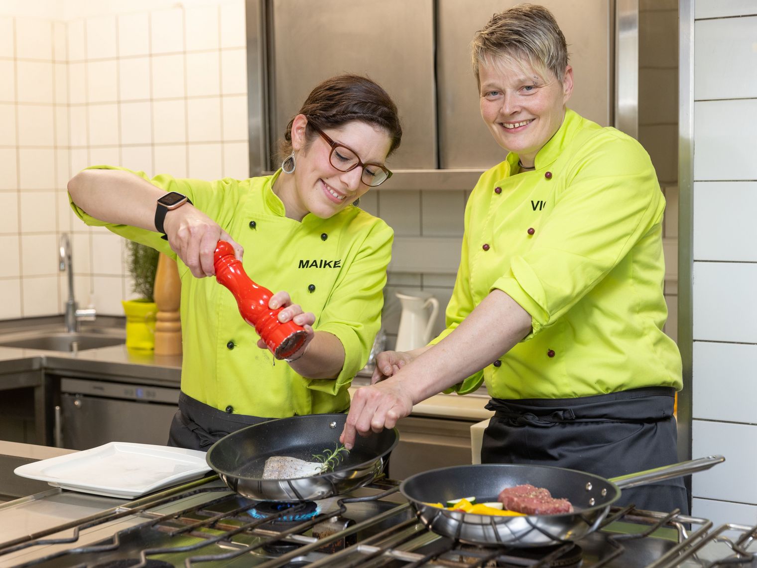Das Bild zeigt zwei Köchinnen in hellgrüner Arbeitskleidung am Herd einer Restaurantküche. 