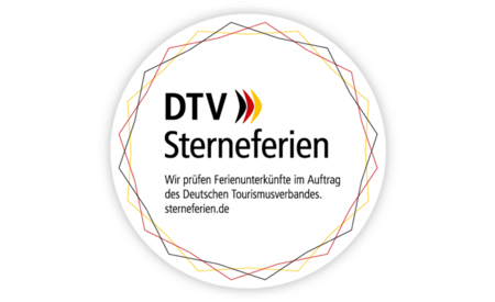 Das Bild zeigt ein rundes Logo mit dem Schriftzug „DTV Sterneferien“ in schwarzen Buchstaben. Daneben befindet sich ein stilisiertes Symbol in den Farben Schwarz, Rot und Gold, das an ein Qualitätssiegel oder eine nationale Kennzeichnung erinnert. Der Rand des Logos ist mit kleinen farbigen Punkten verziert, die dem Design einen freundlichen und einladenden Charakter verleihen.